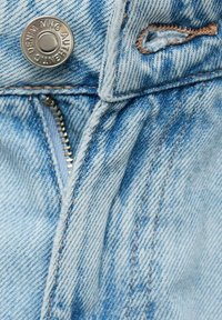 Svjetloplavi traper s vidljivom teksturom, mjedenim zatvaračem i metalnom dugmadi s ugraviranim imenom branda. Šavovi u kontrastnoj boji smeđi.