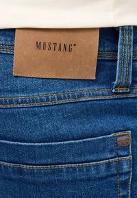 Vaqueros de denim en un tono azul oscuro, con un parche de cuero marrón que lleva la etiqueta "MUSTANG" y un pequeño logo metálico en el bolsillo trasero.