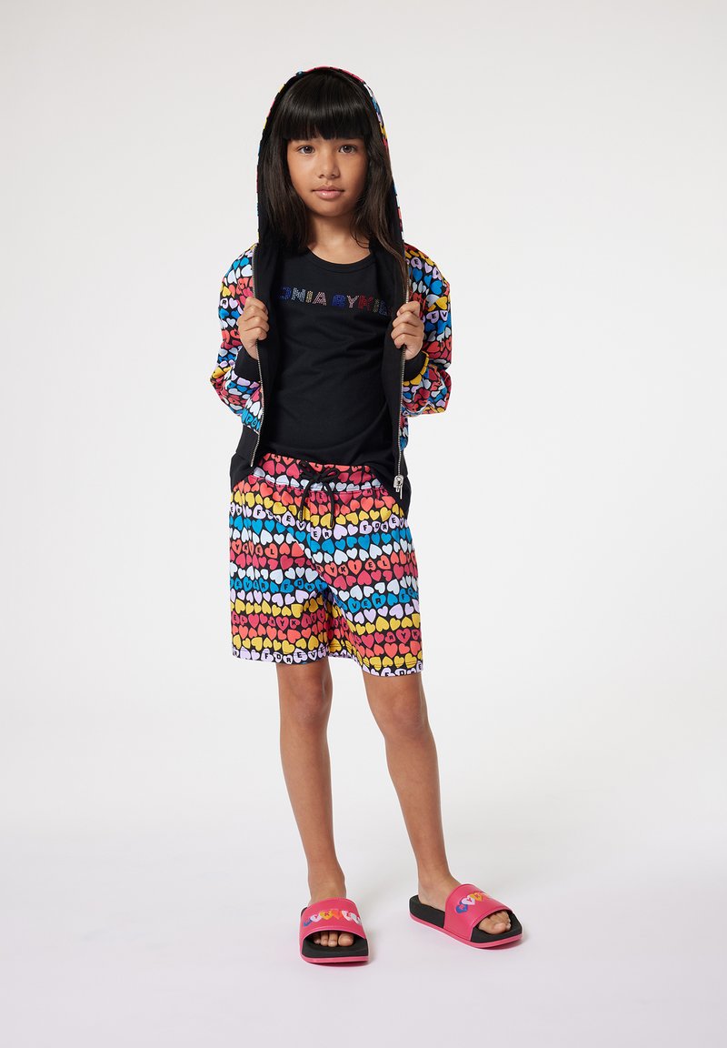 Haut à capuche coloré avec un motif de cœur, t-shirt noir avec logo, shorts assortis et sandales rose/noir. Le matériau semble doux ; le design est ludique.