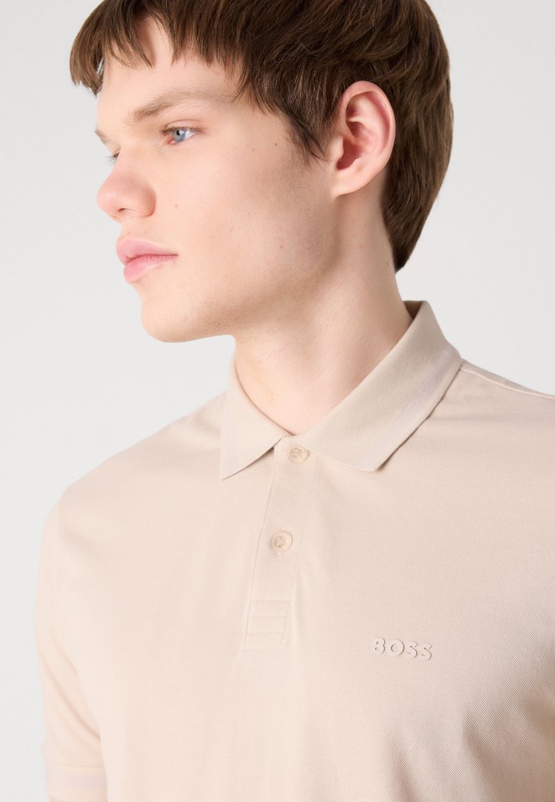 Polo beige avec un col classique, des manches courtes et une patte de boutonnage à trois boutons. Présente un tissu texturé et un logo en relief sur la poitrine.