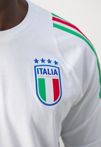 Bílý sportovní triko s modrými a zelenými pruhy, vyšitým odznakem Italia s červenými, bílými a zelenými prvky a pěti modrými hvězdami.