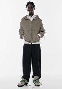Beige pletená mikina na zip s velkým límcem, která je kombinována s bílou košilí a volnými černými kalhotami. Celkový vzhled doplňují šedé a zelené šněrovací boty.