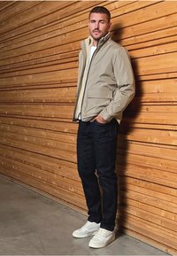 Veste légère beige avec un col montant, poches à l'avant, portée sur une chemise blanche. Associée à un jean noir et des baskets blanches. Fond en panneaux de bois.