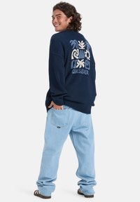 Pulôver de malha azul-marinho com um gráfico em creme estampado nas costas, combinado com jeans baggy azul-claro e sandálias castanhas.