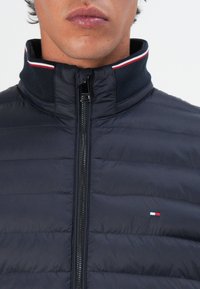 Marineblå puffjakke med høy krage, horisontal quiltet tekstur, frontglidelås og liten logo med røde, hvite og blå detaljer.