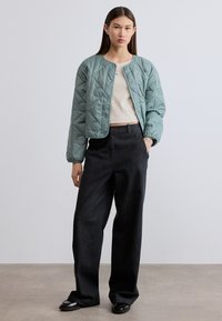 Πράσινο πουπουλένιο μπουφάν με στρογγυλή λαιμόκοψη και κουμπιά με πατιλέτα, σε συνδυασμό με μπεζ τοπ και σκούρο τζιν παντελόνι με φαρδιά μπατζάκια. Μαύρα παπούτσια προσθέτουν μια κομψή πινελιά.