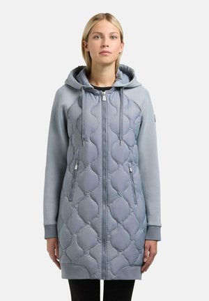 Kobieta ubrana w długą, jasnoniebieską pikowaną kurtkę z kapturem, zapinaną na zamek błyskawiczny z przodu i z zamykanymi na zamek kieszeniami, stojąca na jednolitym tle.