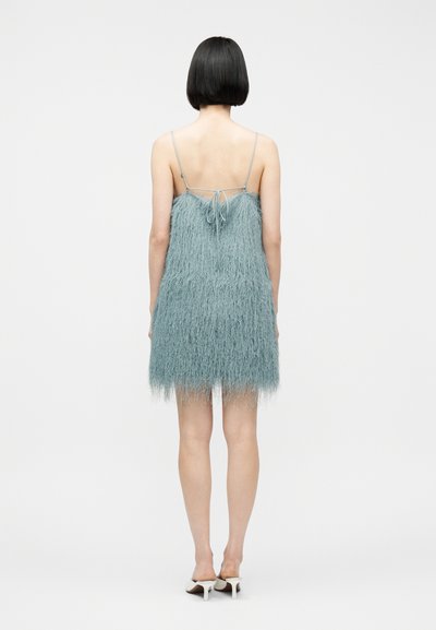 Γυναίκα με κοντά μαύρα μαλλιά καρέ, που φοράει αμάνικο, μπλε υφασμάτινο μίνι φόρεμα με λεπτές τιράντες και λευκά σανδάλια με τακούνια, στέκεται με την πλάτη προς το μέρος μας.