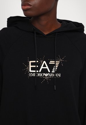 Personne à la peau foncée portant un sweat à capuche noir avec le logo "EA7 Emporio Armani" et des paillettes décoratives sur la poitrine.