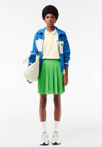 Giacca blu lavorata a maglia con dettagli bianchi, polo color crema e gonna plissettata verde. Borsa a tracolla bianca e sneakers sportive completano l'outfit.
