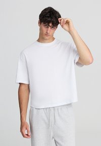 Biała koszulka z krótkim rękawem o luźnym kroju i okrąg�łym dekoltem, charakteryzująca się gładką fakturą, noszona z szarymi spodniami dresowymi.