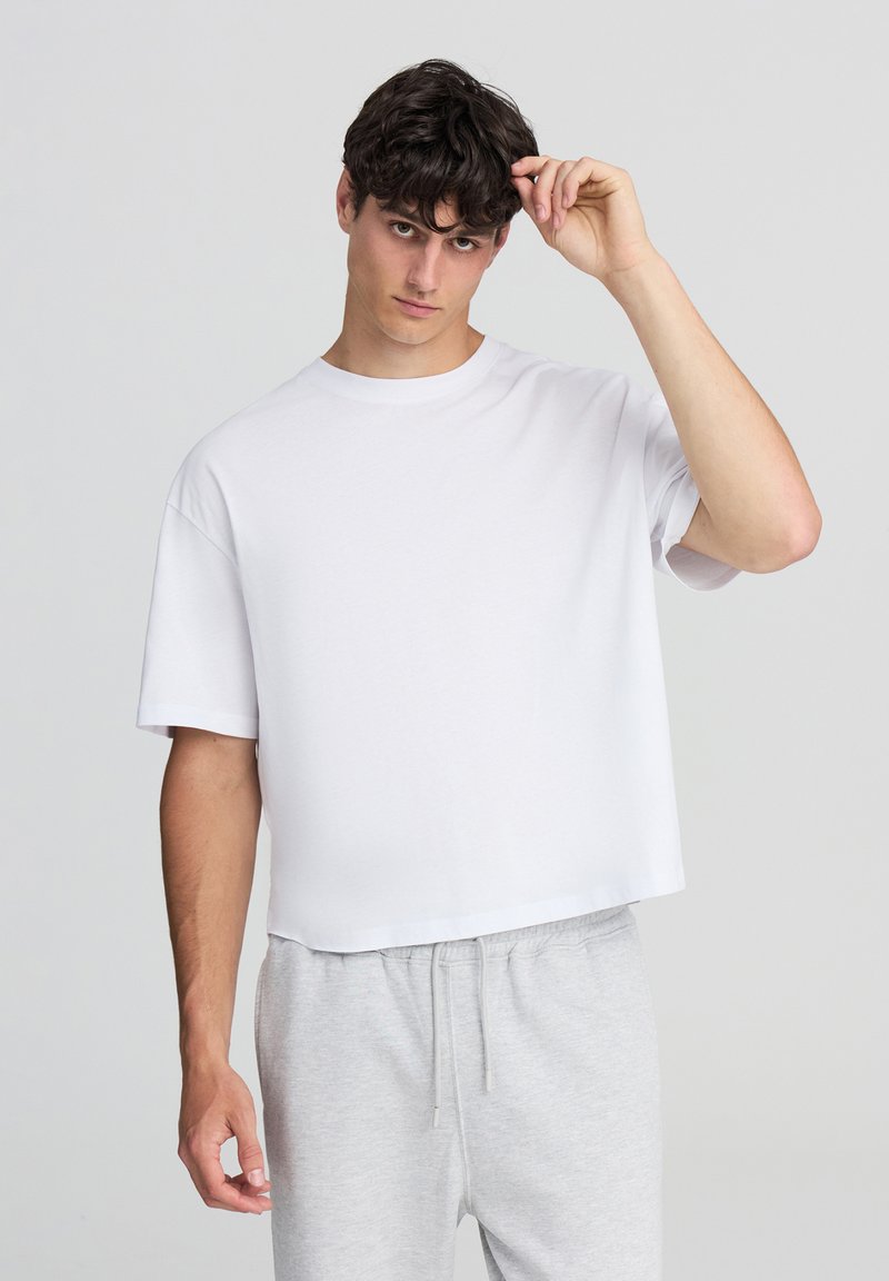 Biała koszulka z krótkim rękawem o luźnym kroju i okrągłym dekoltem, charakteryzująca się gładką fakturą, noszona z szarymi spodniami dresowymi.