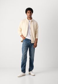 Giacca button-up crema chiaro sopra una maglietta bianca, abbinata a jeans di denim blu e sneakers bianche, in piedi su uno sfondo semplice.