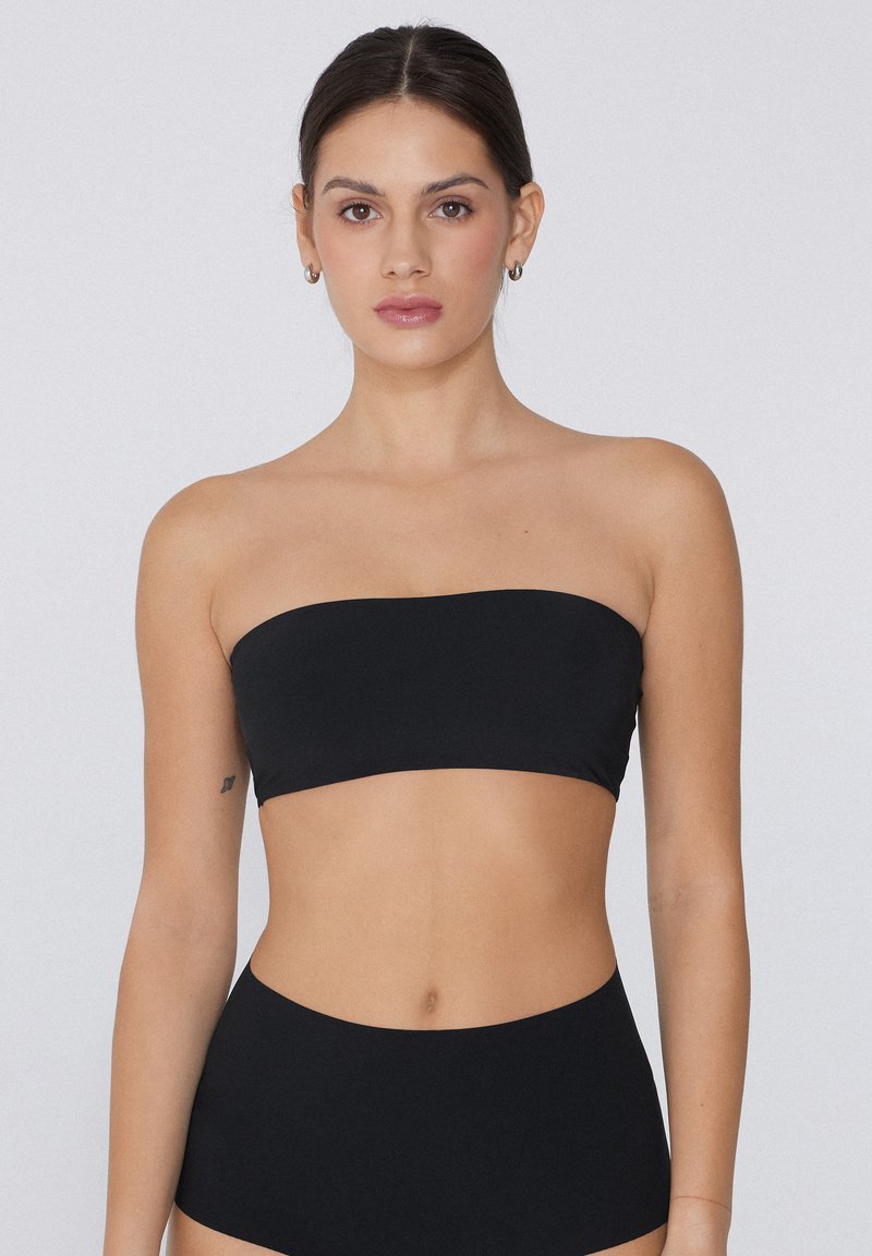Joven con cabello oscuro con un top bandeau negro sin tirantes y pantalones de talle alto contra un fondo claro y liso.