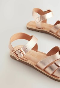 Sandalias metálicas en oro rosa con tiras cruzadas de textura suave, suela marrón y correa ajustable en el tobillo, que muestran un diseño simple y abierto.