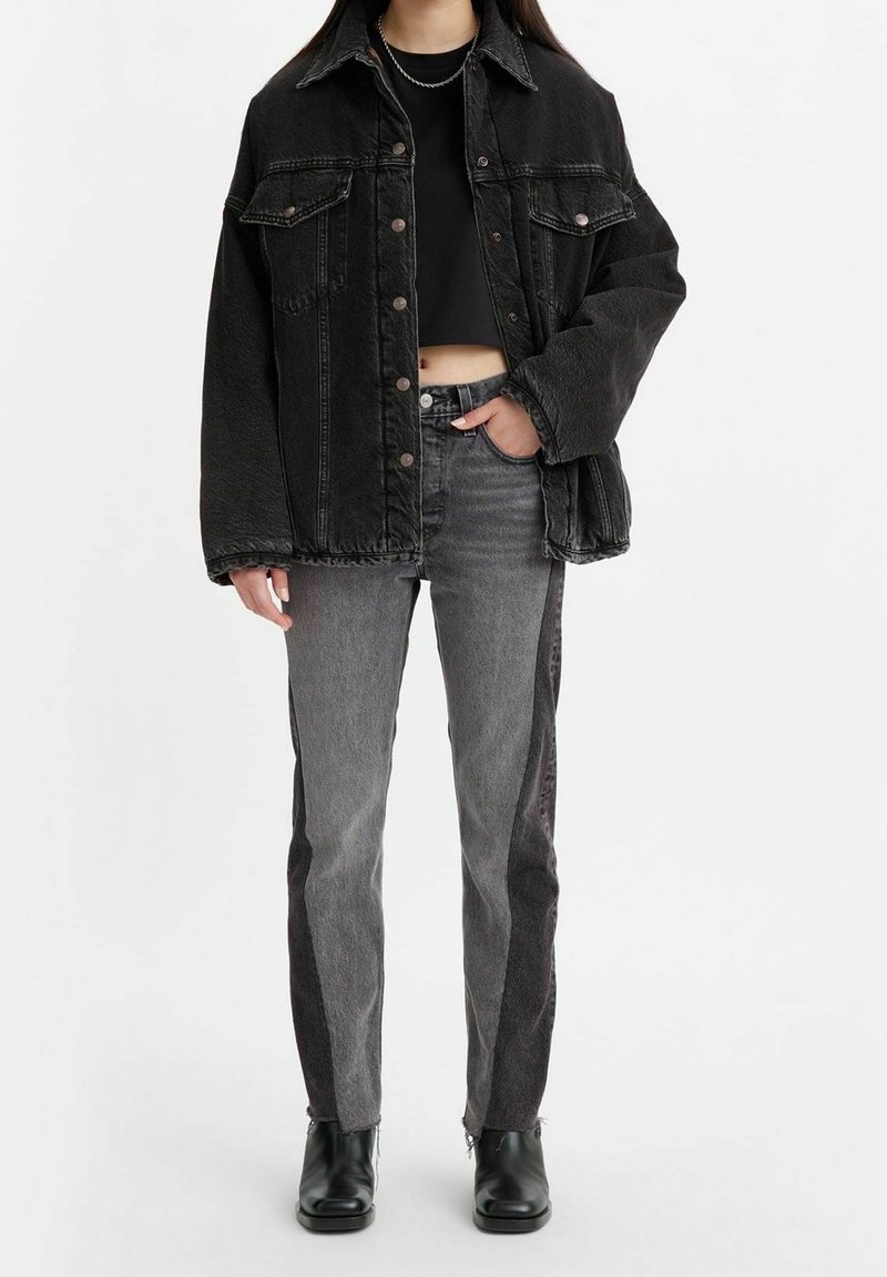 Personne portant une veste en jean noire surdimensionnée, un crop top noir, un jean bicolore gris et des bottines noires, debout avec une main dans la poche.