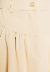 Ljusbeige kjol i mjukt tyg, med en draperad detalj, en slät yta och en enda rund knapp i midjan.