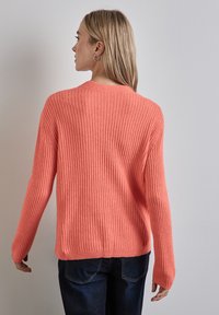 Koralfarbener Ripppullover mit langen Ärmeln, lockerer Passform und rundem Ausschnitt. Weicher Stoff und ein Saum, der auf Taillenhöhe endet.