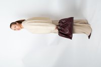Femme se tenant debout dans un pantalon large beige et un pull en crème pelucheux, tenant un grand sac à main en cuir bourgogne foncé contre un fond blanc.