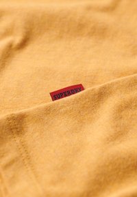 Tela de algodón amarilla con una textura fina, que presenta una etiqueta roja con "SUPERDRY" impresos en negro. La tela está dispuesto de manera suave.