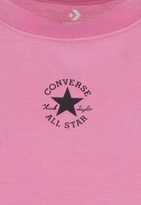 Roosa T-särk, millel on must ümmargune "Converse All Star" logo keskel oleva tähe ja tekstiga. Valmistatud pehmest puuvillast kangast.