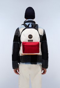 Le sac à dos possède un corps principal blanc, une poche avant rouge et des accents noirs. Il est porté par-dessus une veste en polaire à carreaux avec des motifs bleus et noirs.