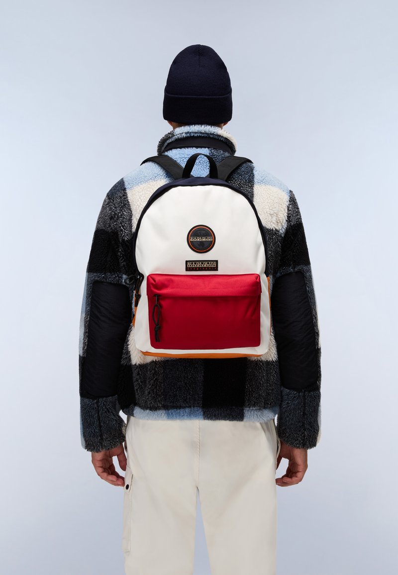Le sac à dos possède un corps principal blanc, une poche avant rouge et des accents noirs. Il est porté par-dessus une veste en polaire à carreaux avec des motifs bleus et noirs.
