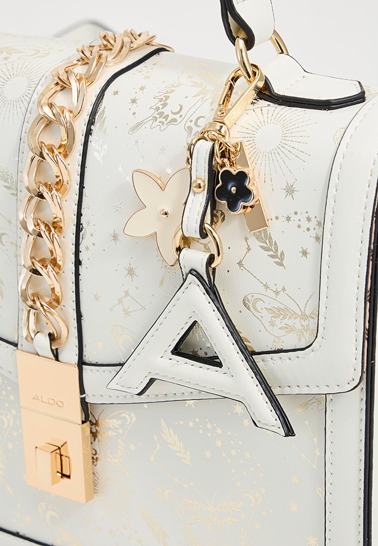 Sac à main en cuir blanc avec motif floral doré, une chaîne chunky en or, et des charms décoratifs comprenant une fleur et la lettre "A".