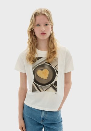 Witte katoen t-shirt met een grafische print van een hartvormige pannenkoek in een zwarte koekenpan, met een eenvoudige ronde halslijn en korte mouwen.