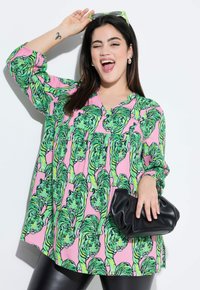 Lyserød skjorte med grøn tigerprint, lange ærmer, v-hals; kombineret med en sort clutch og blanke sorte leggings. Glat stoftekstur.