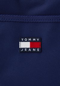 Tengerészkék szövetjelvény, rajta a "TOMMY JEANS" felirat egy vízszintes piros, fehér és kék csíkos mintázat felett, textilfelületre varrva.