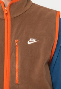 Hnědá fleecová vesta s oranžovým zipem a akcenty, má náprsní kapsu a bílé logo Nike na přední straně. Hladký povrch, vysoký límec.