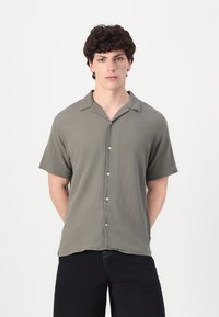 Camisa de manga corta abotonada en color verde oliva, con tejido texturizado, que presenta un cuello tipo campamento y un cierre frontal de cinco botones. Pantalones negros visibles.
