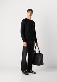 Schwarzer Pullover mit langen Ärmeln und kleinem Logo, kombiniert mit dunklen Jeans und schwarzen Lederschuhen. Hält eine schwarze Ledertasche.