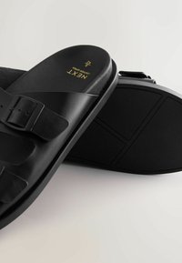 Sandales à enfiler en cuir noir avec sangles à double boucle, une sandale montrant la semelle texturée, taille 42 avec une étiquette dorée "NEXT Leather Upper" à l'intérieur.