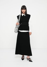 Black layered outfit featuring a sleeveless knit vest over a white shirt, paired with a long black skirt, black heels, and a patterned handbag.