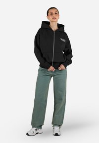 Felpa nera con zip, dotata di una grande tasca frontale e logo. Abbinata a jeans ampi verde chiaro e sneakers bianche. Tessuto dalla superficie liscia.