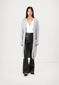Hellgrauer, langer Cardigan aus weichem Strickmaterial, mit gerippten Säumen und Taschen. Kombiniert mit engen schwarzen Lederhosen und Clogs.