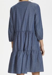 Žena oblečená v modrých šatech po kolena s tříčtvrtečními rukávy a jemným vzorem listů, zobrazená zezadu.