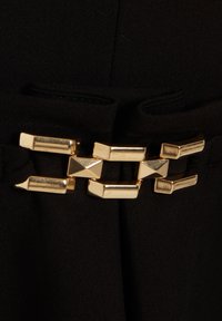 Tissu noir avec une boucle géométrique dorée comprenant des formes rectangulaires et pyramidales. La boucle est intégrée au design du vêtement.