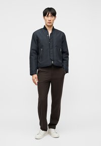 Άνδρας που στέκεται φορώντας μαύρο μπουφάν τύπου bomber, σκούρο καφέ παντελόνι και λευκά αθλητικά παπούτσια σε απλό λευκό φόντο.