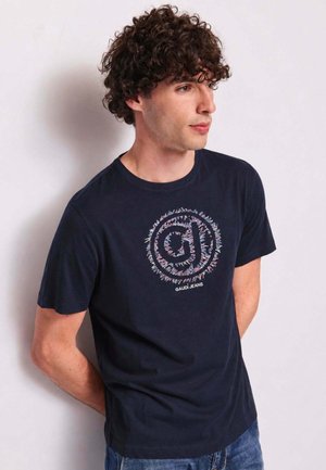 Uomo con capelli ricci che indossa una maglietta Gaudi Jeans blu scuro, guarda di lato con le mani dietro la schiena contro un muro semplice.