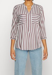 Blusa grigia chiaro con righe verticali bordeaux, maniche a tre quarti, scollo a V e due tasche sul petto, realizzata in tessuto morbido.