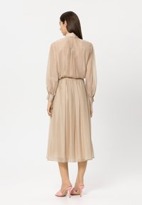 Beige long-sleeve dress with a gathered waist and pleated skirt. Sheer fabric with subtle texture. Worn with pink high-heeled shoes.