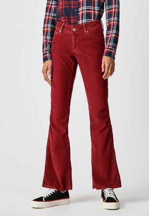 Persona con pantalones acampanados de pana roja, camisa de cuadros azul marino y rojo con botones, y zapatillas negras con suelas y cordones blancos, de pie sobre fondo blanco.