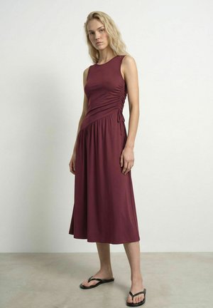 Femme blonde portant une robe midi bordeaux sans manches avec fronces latérales et des tongs noires, debout contre un mur clair uni.