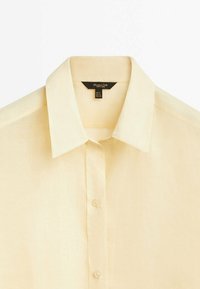 Chemise jaune pâle à manches courtes avec un col classique, une patte de boutonnage à l'avant et un tissu au toucher doux. L'étiquette indique la taille et la marque.