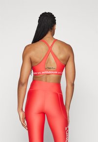 Red sports bra with crisscross straps and Under Armour logo. Paired with high-waisted red leggings, smooth texture, form-fitting design.