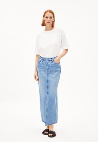 Witte t-shirt met korte mouwen gecombineerd met een hoog opgestelde lichtblauwe denim maxi-rok met een rechte pasvorm. Model draagt zwarte instap sandalen.