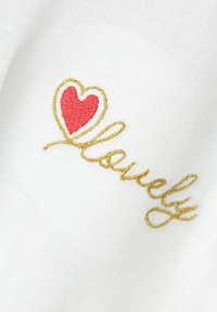 Tissu blanc avec le mot brodé "lovely" en fil doré, accentué par un cœur rouge. Texture lisse et design épuré.